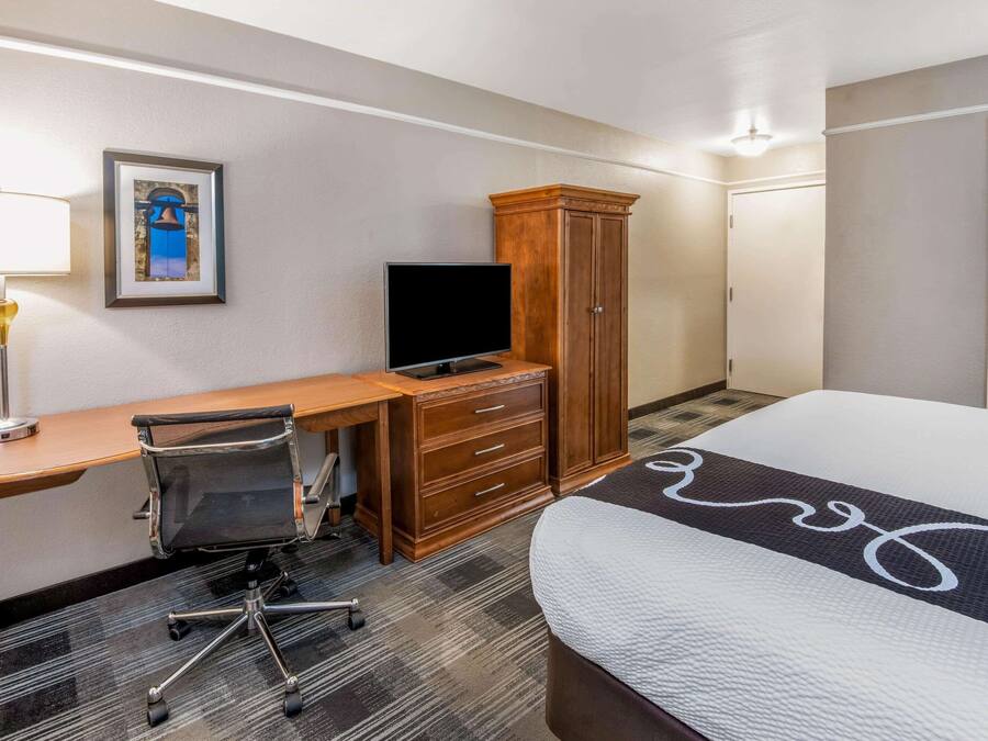 A hotel room with a bed, a desk with a chair, a television, a wardrobe, and a framed picture on the wall.