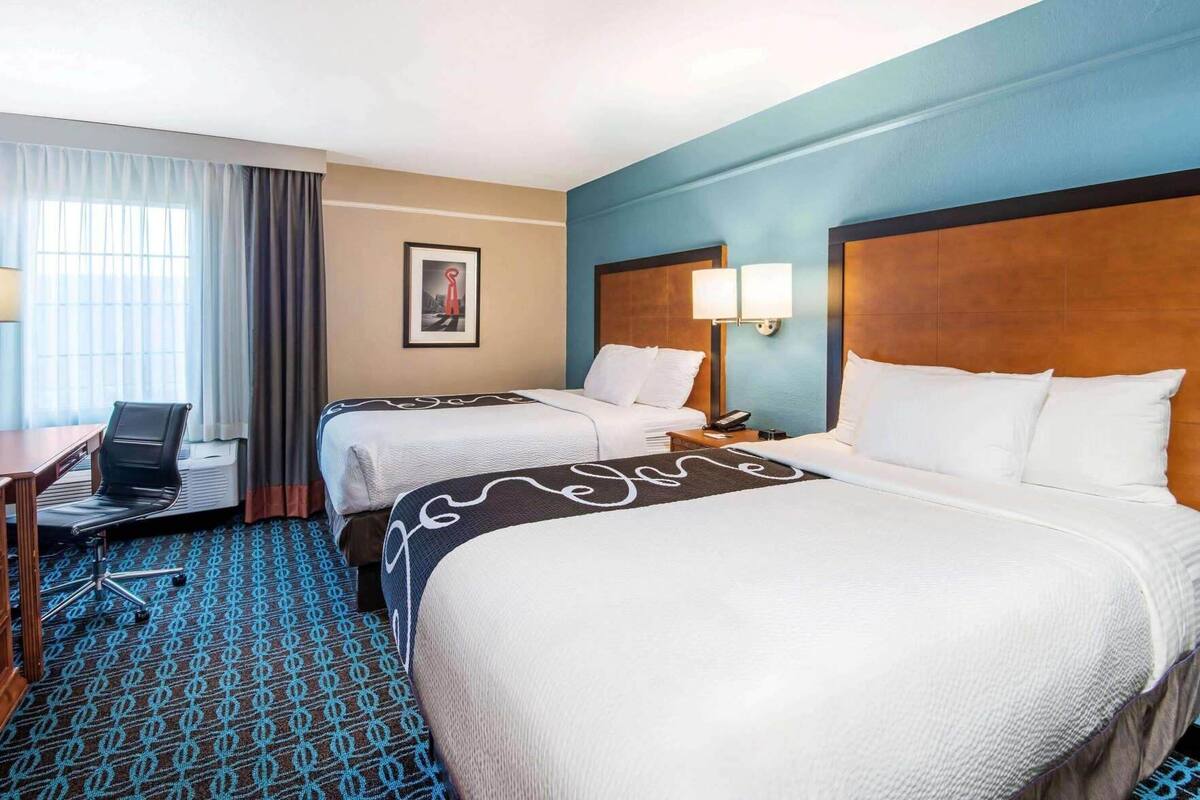 A hotel room with two beds, a desk with a chair, a lamp, and a framed picture on the wall.
