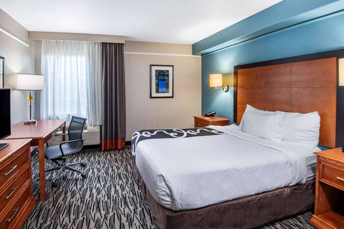 A hotel room with a bed, desk, chair, and a view of the outdoors through a window.