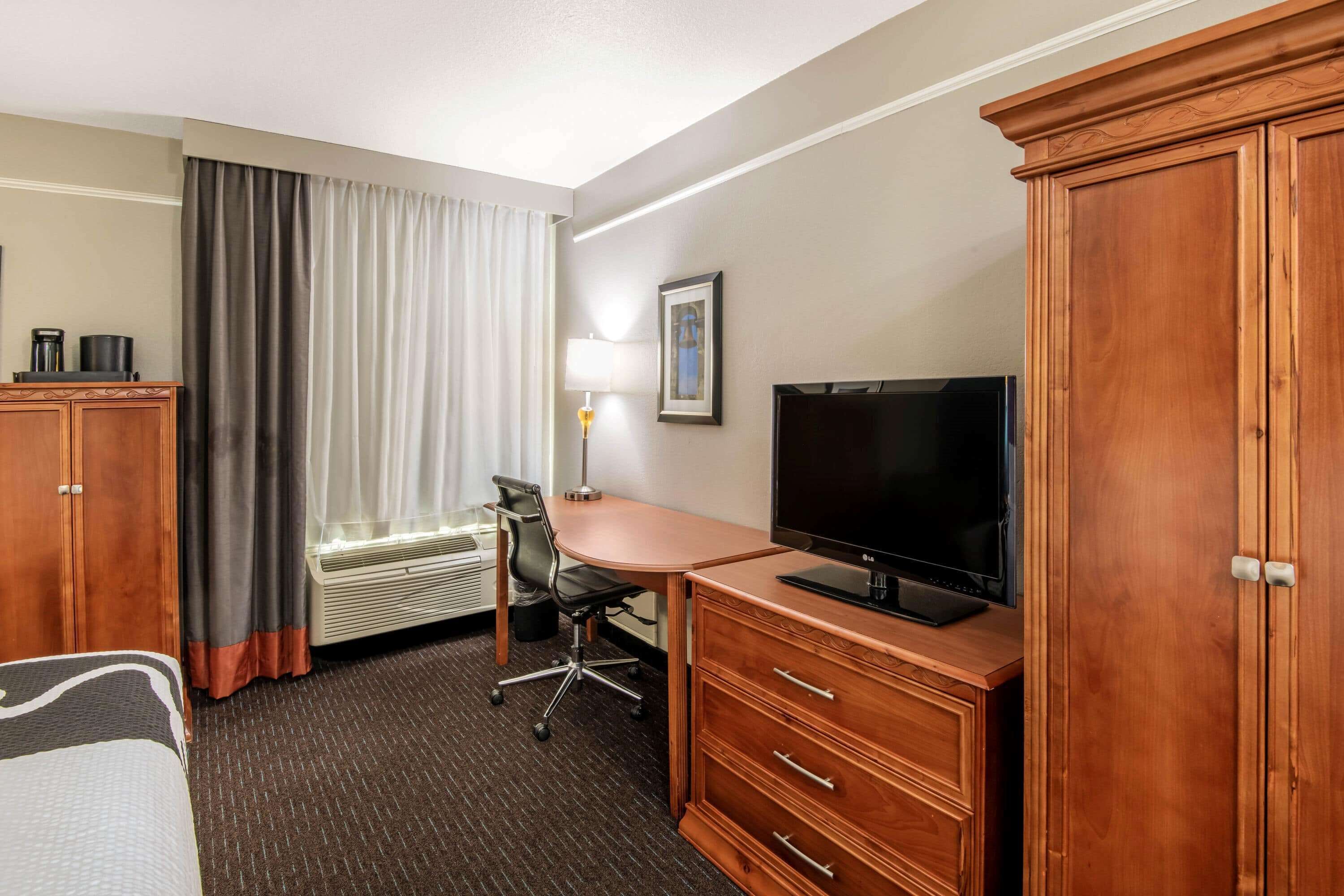 Habitación de hotel con cama, escritorio con televisor, silla, armario y ventana con cortinas.