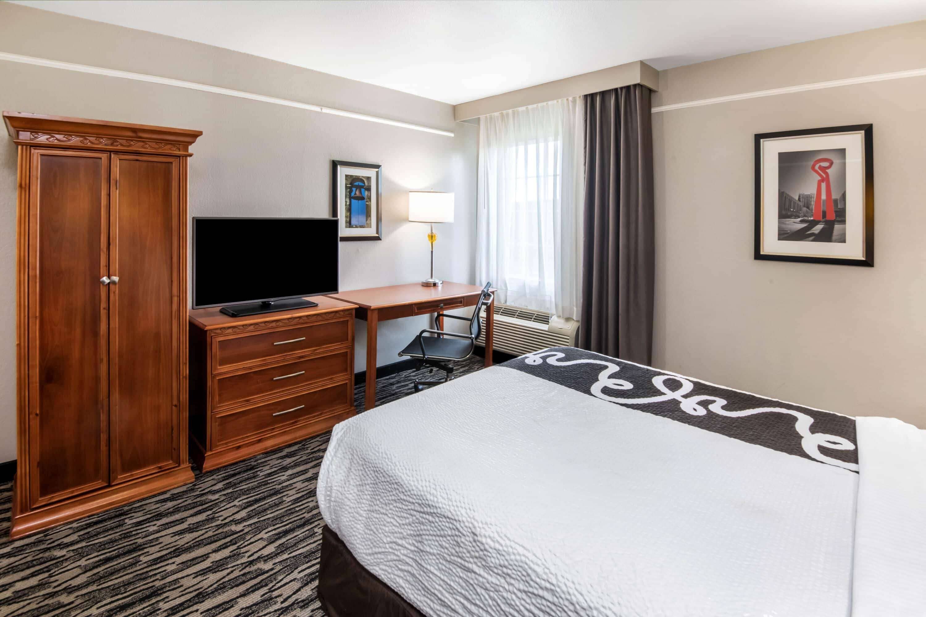 Habitación de hotel con cama, escritorio, televisor, armario y ventana con cortinas.