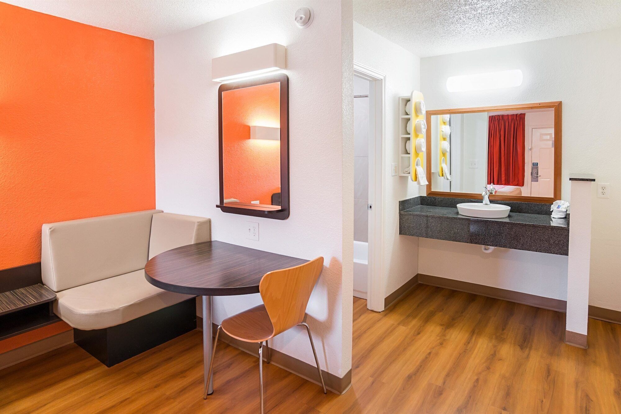 A hotel room with a wooden floor, a round table with a chair, a sofa, a bathroom with a sink and mirror, and a red curtain.