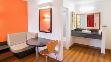A hotel room with a wooden floor, a round table with a chair, a sofa, a bathroom with a sink and mirror, and a red curtain.
