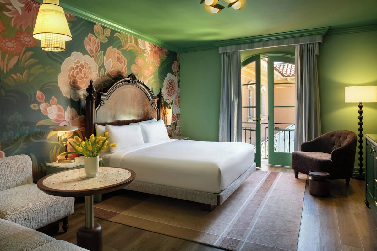 A hotel room with a bed, a chair, a small table, a lamp, and a floral wall mural.