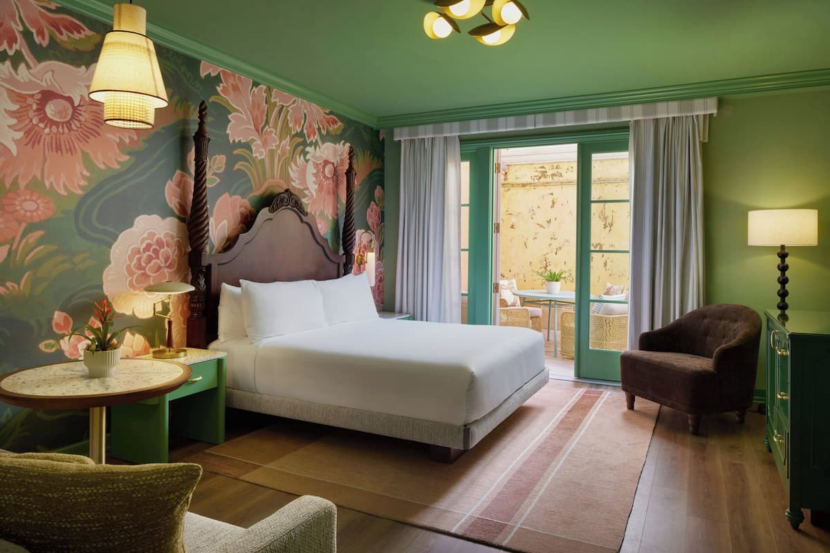 A hotel room with a bed, a chair, a small table, a lamp, and a view of the outside.