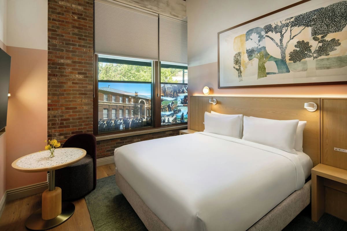 A hotel room with a large bed, a small round table with a vase, a window with a view of buildings, and a painting on the wall.