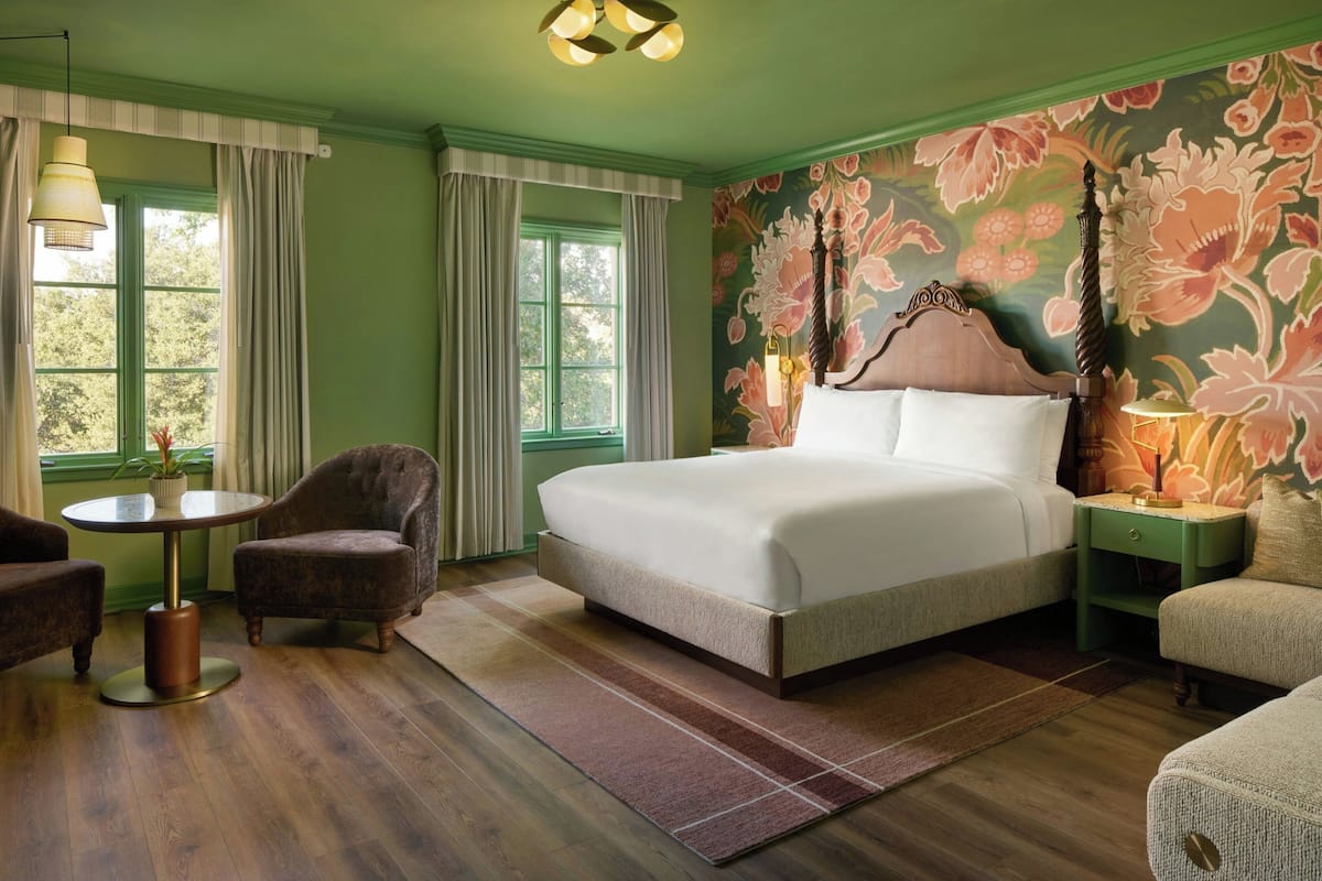 A bedroom with a large bed, a round table, two armchairs, a nightstand, and a floral wallpaper.