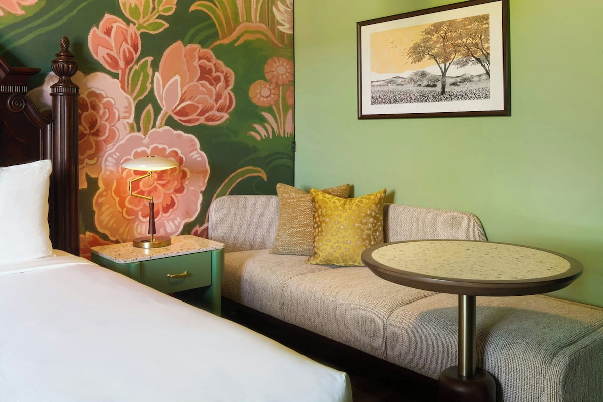 A room with a bed, a nightstand with a lamp, a sofa with cushions, a small table, and a framed picture on the wall.