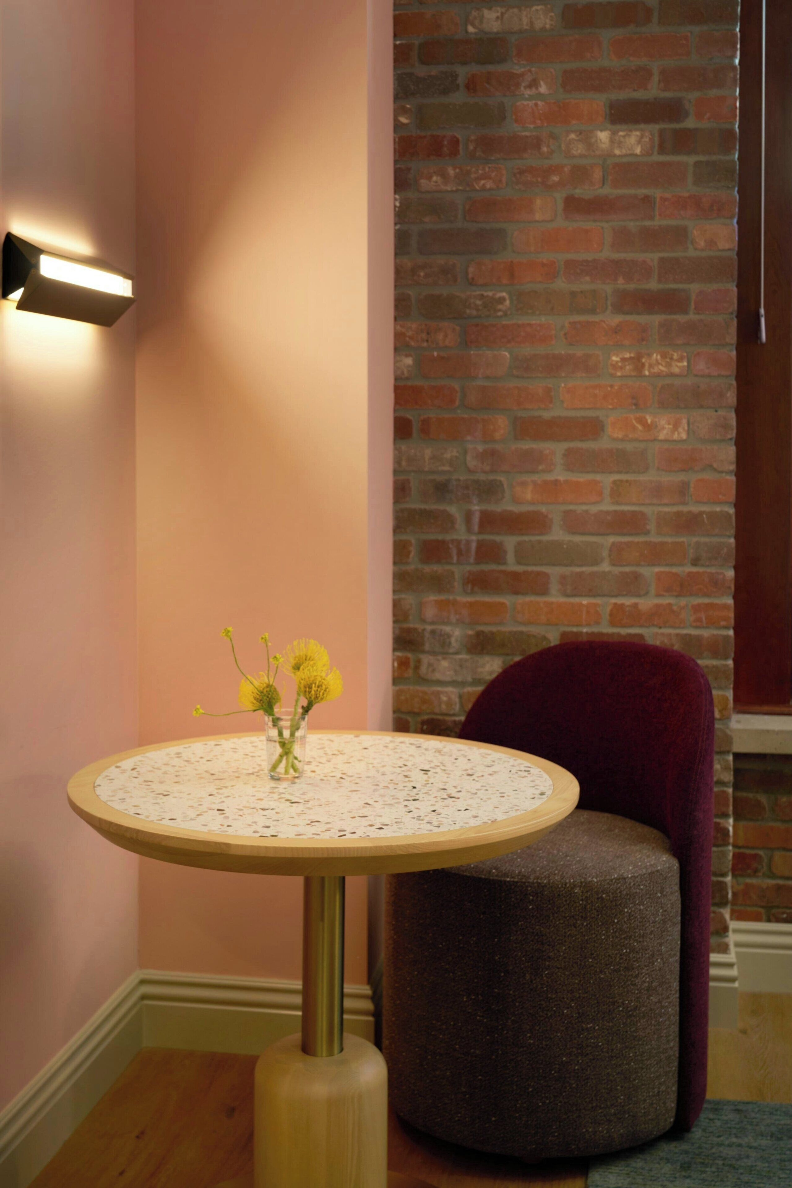 A round table with a vase of yellow flowers, a brick wall, and a red chair in a corner.