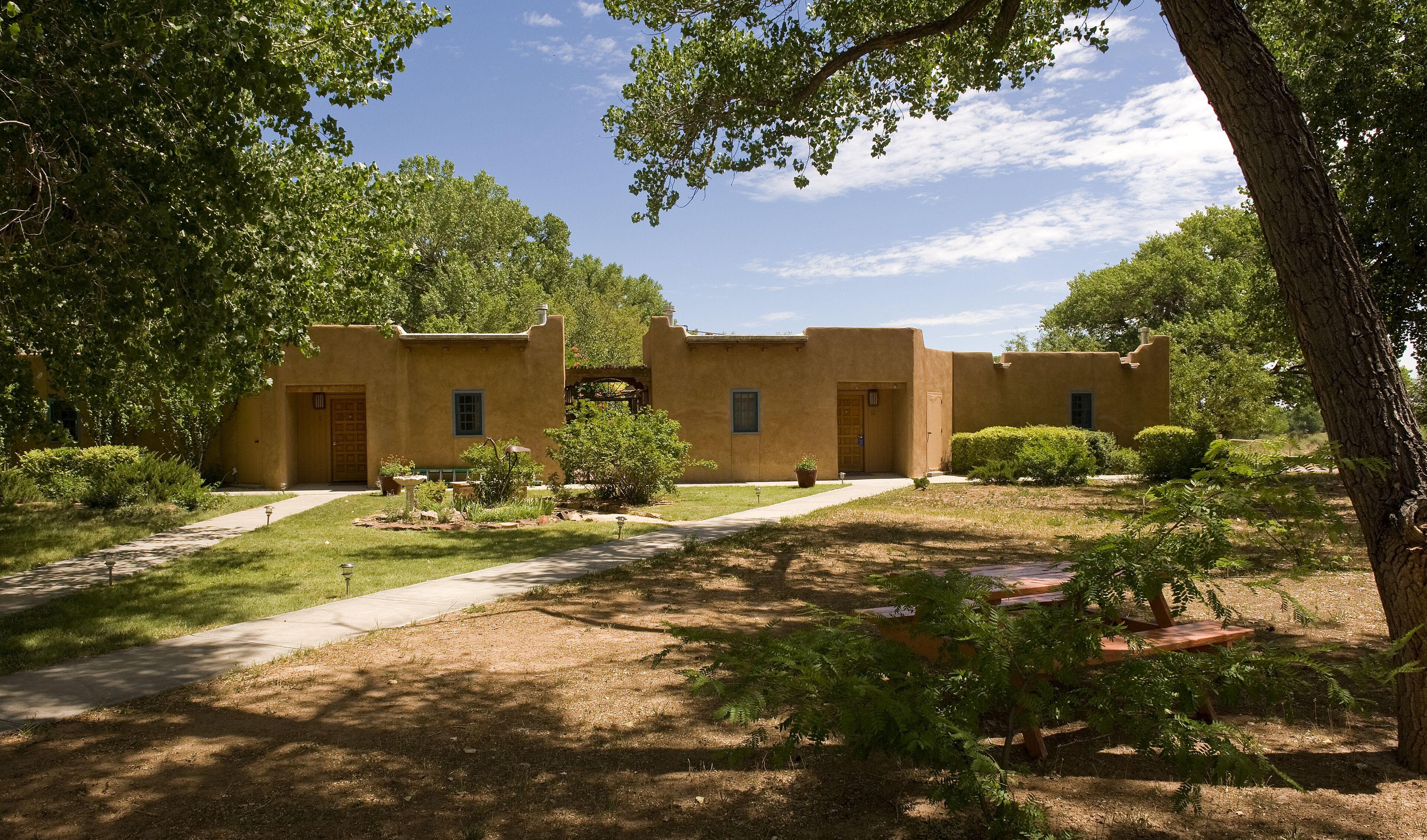 A courtyard with a building featuring a flat roof and stucco walls, surrounded by trees and a paved walkway.