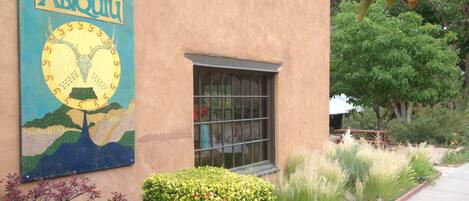 Un edificio con un mural de un árbol y montañas, una ventana, y un jardín bien cuidado con diversas plantas y flores.
