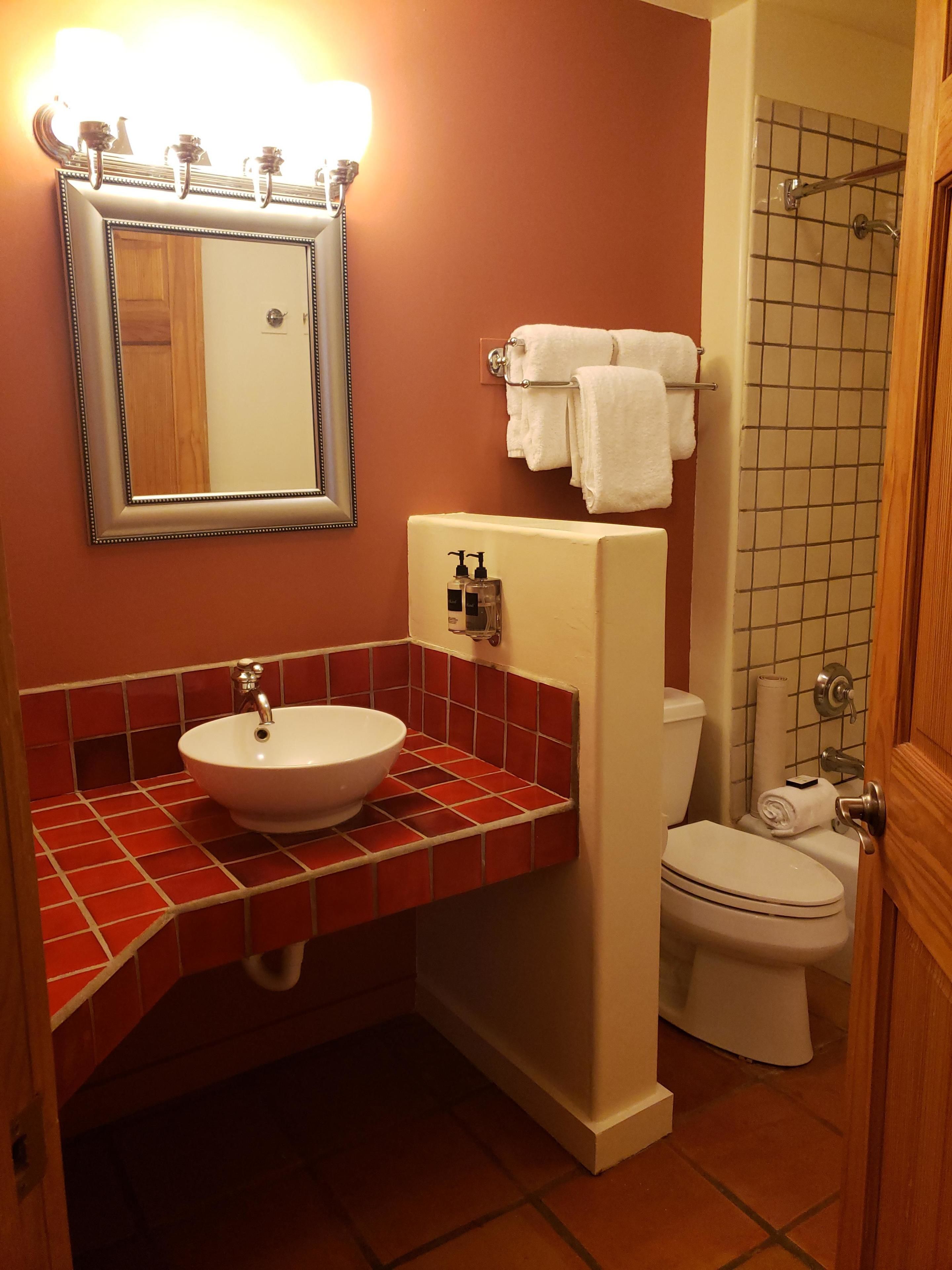 A bathroom with a white sink, red-tiled vanity, a mirror with lights, a toilet, and a shower area.