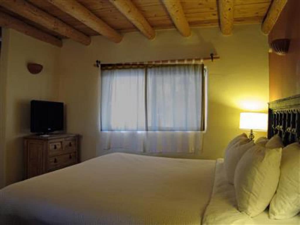 A bedroom with a bed, a television on a wooden dresser, a window with curtains, and a lamp on the nightstand.