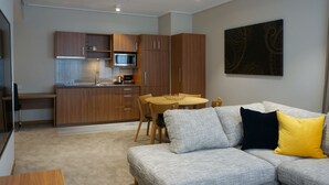 Una cocina moderna con armarios de madera y un microondas, un área de comedor con una mesa redonda y sillas, y un sofá gris con cojines amarillos y negros.