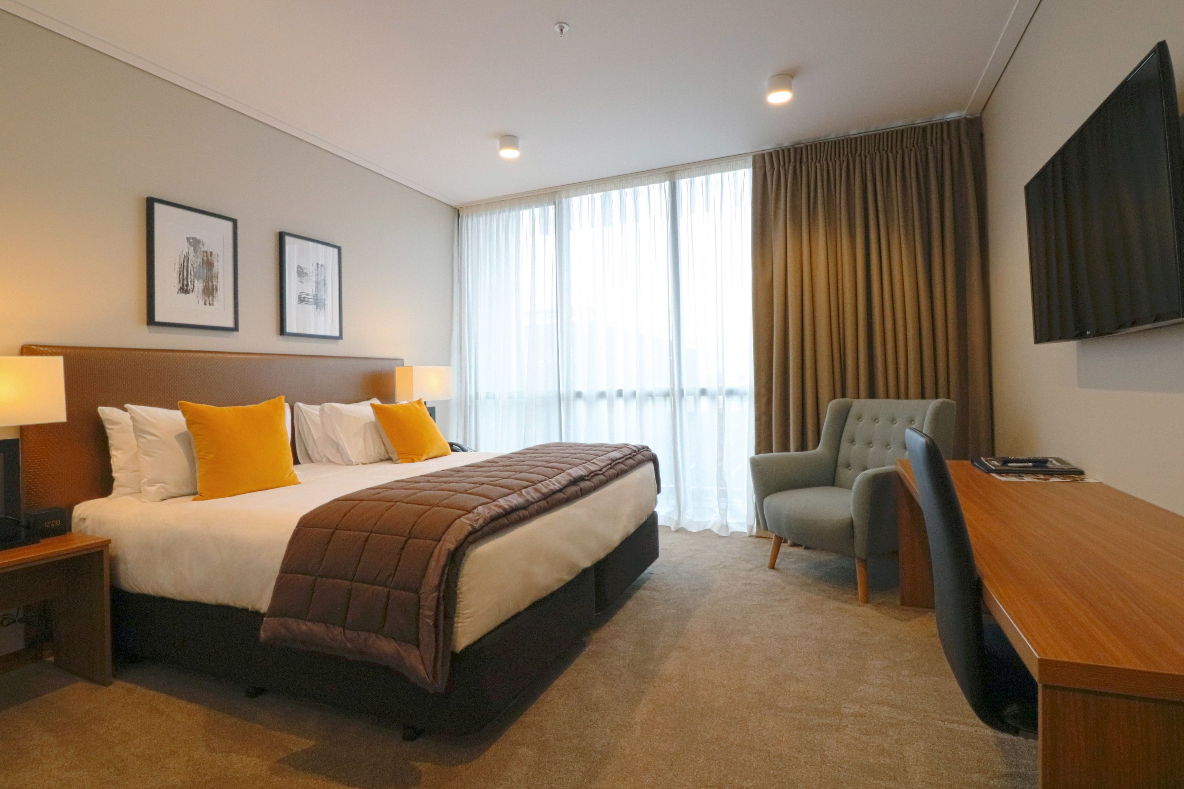 Habitación de hotel con una cama grande, un escritorio, una silla, un televisor y dos cuadros en la pared.