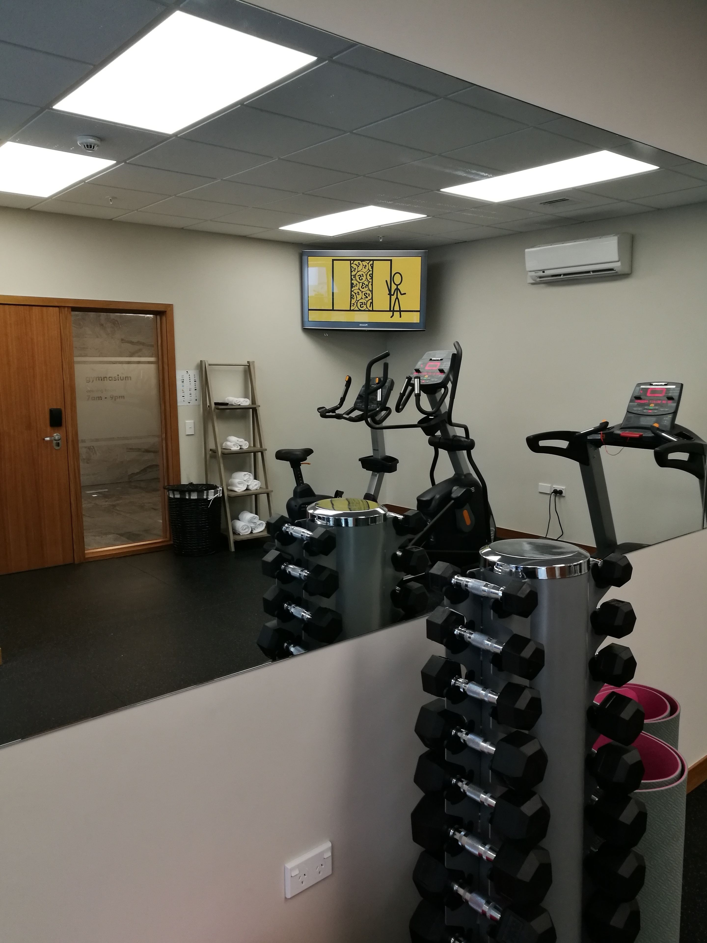 Une salle de sport équipée de matériel d’exercice, d’un tapis de course et d’un téléviseur fixé au mur.