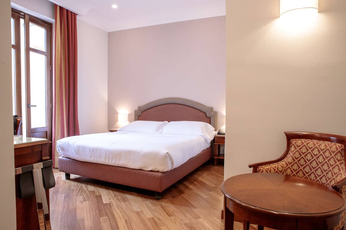 A neatly made bed with a wooden headboard, a wooden chair, and a small table in a room with wooden flooring and a window with curtains.