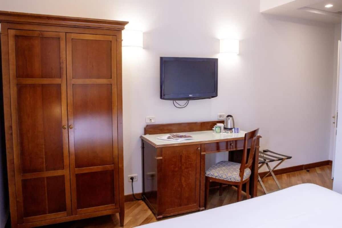 A hotel room with a wooden wardrobe, a desk with a chair, a flat-screen TV mounted on the wall, and a bed with a white duvet.