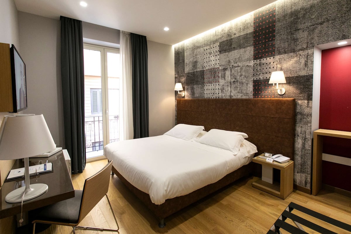 A hotel room with a large bed, a desk with a lamp, a chair, and a view of the outside through a window.