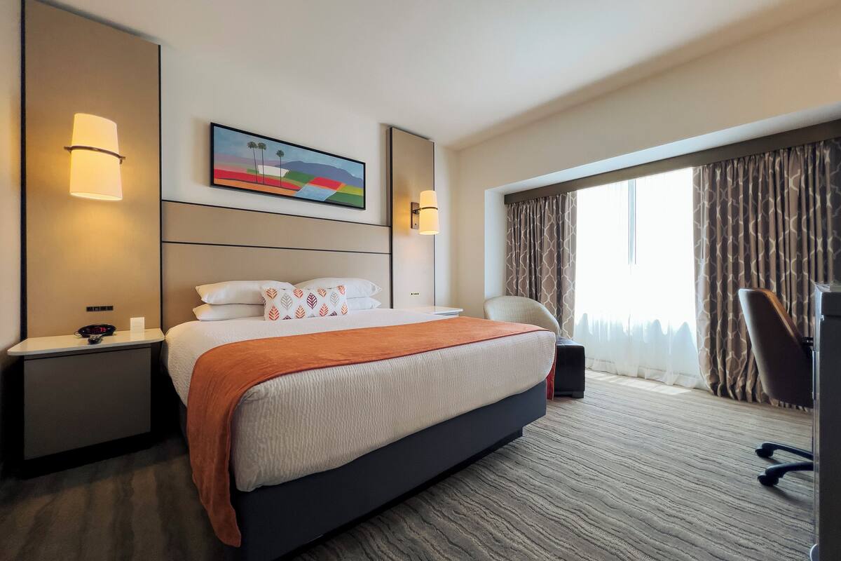 Une chambre d’hôtel avec un grand lit, deux tables de chevet, un bureau, une chaise et une grande fenêtre avec des rideaux.
