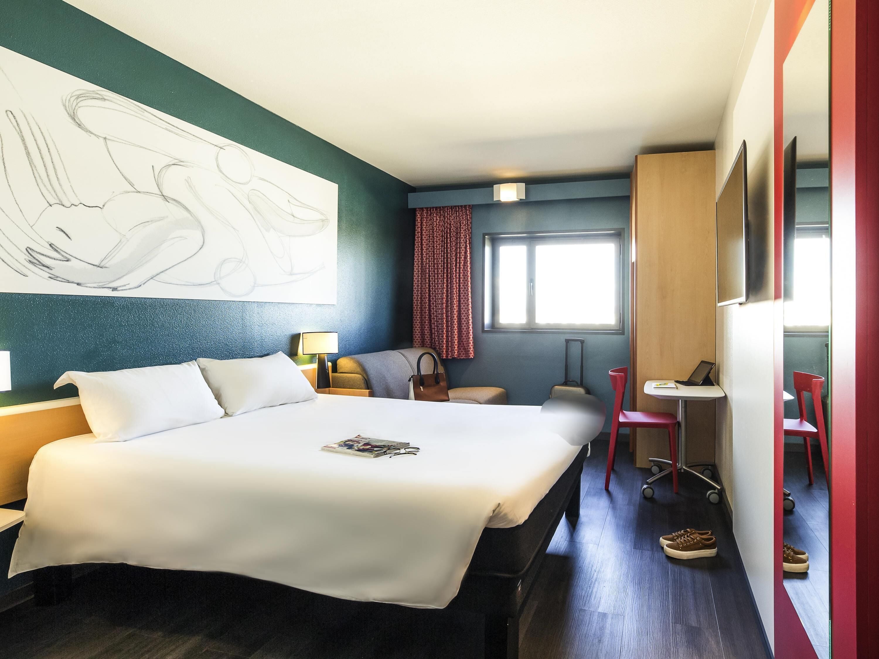 A hotel room with a large bed, a desk, a chair, a suitcase, and a wall art piece.