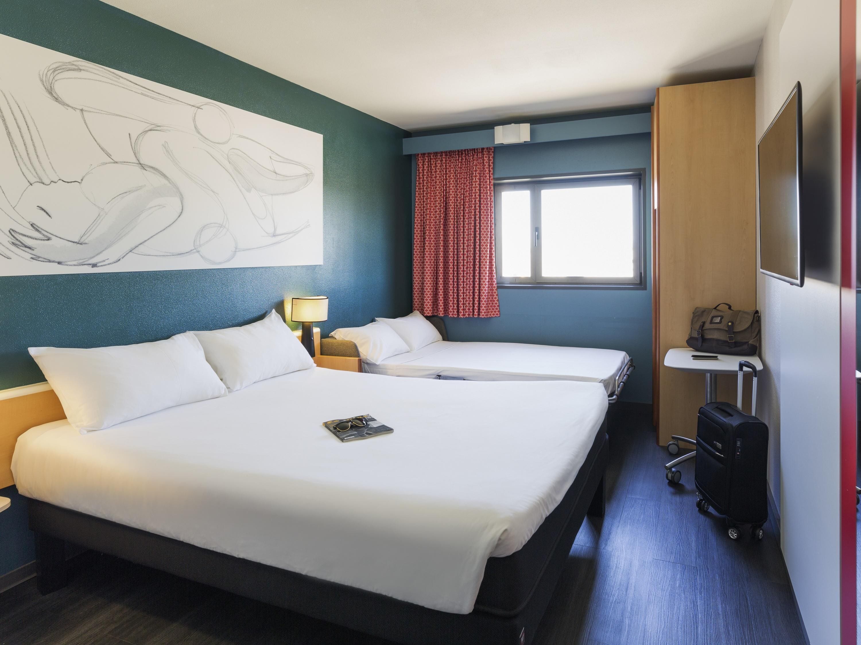 Habitación de hotel con dos camas, un escritorio, una lámpara, una maleta y un cuadro abstracto grande en la pared.