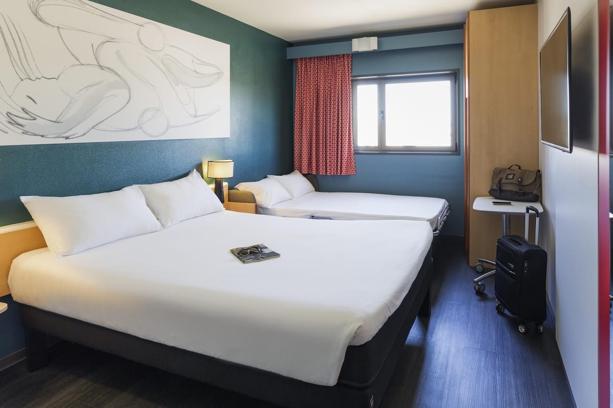 Une chambre d’hôtel avec deux lits, un bureau, une lampe, une valise et un grand tableau abstrait accroché au mur.