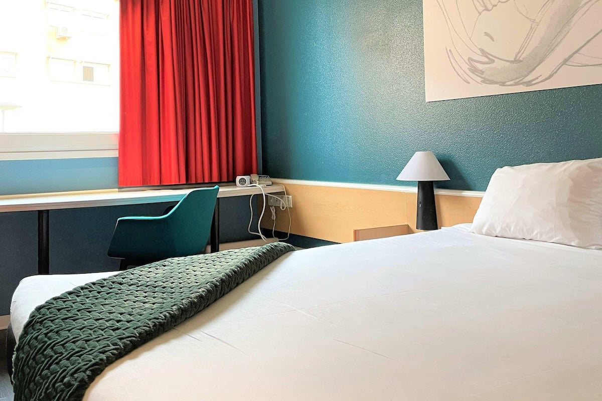Une chambre d’hôtel avec un lit, un bureau, une chaise, une lampe et une fenêtre avec des rideaux rouges.