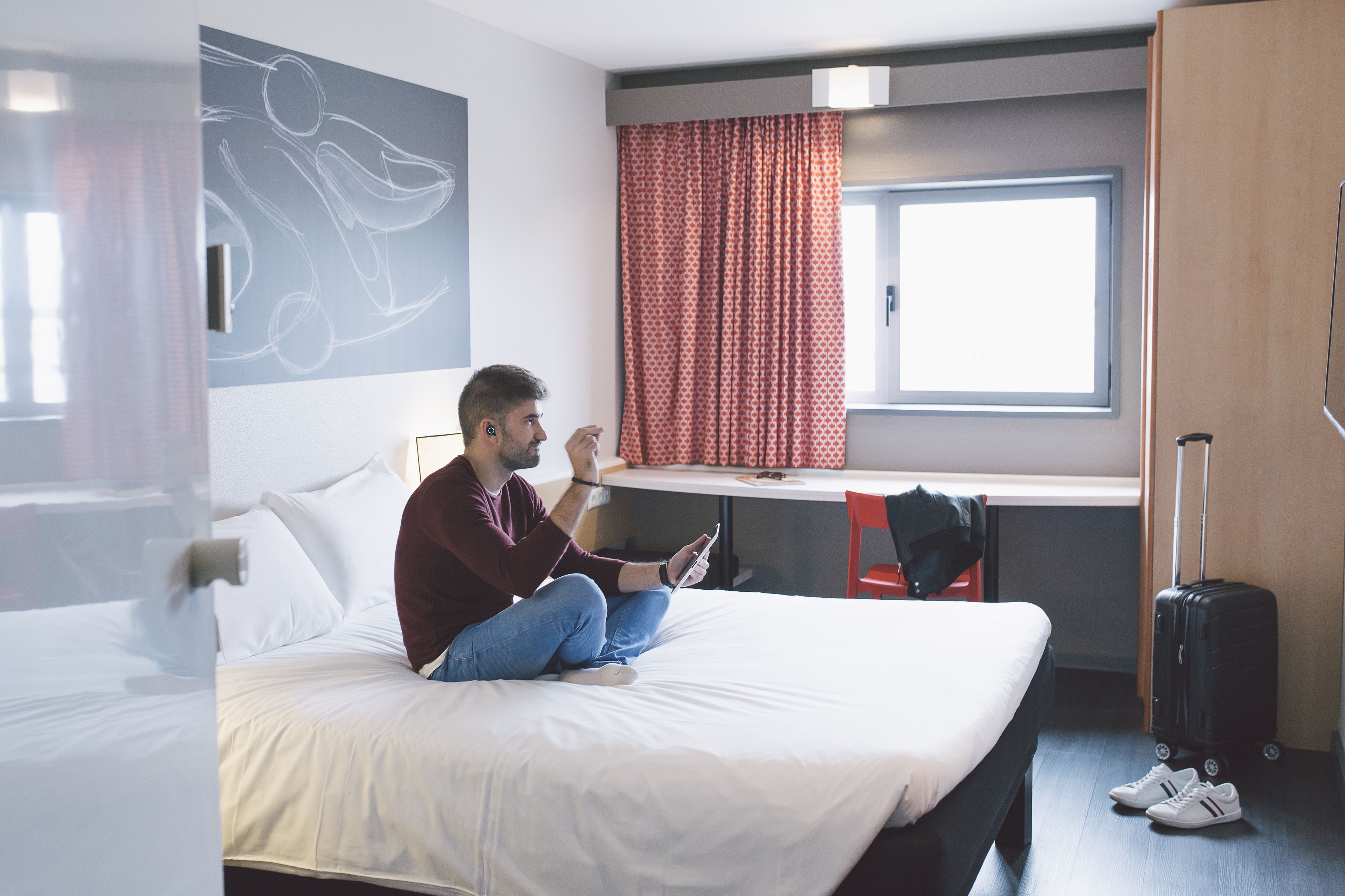 Une chambre d’hôtel avec un lit, un bureau, une fenêtre avec des rideaux, une valise et une paire de chaussures.