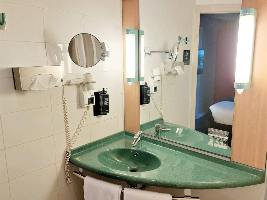 Baño con un lavamanos verde, un espejo, un secador de pelo, un perchero para toallas y un inodoro que se ve en el reflejo.
