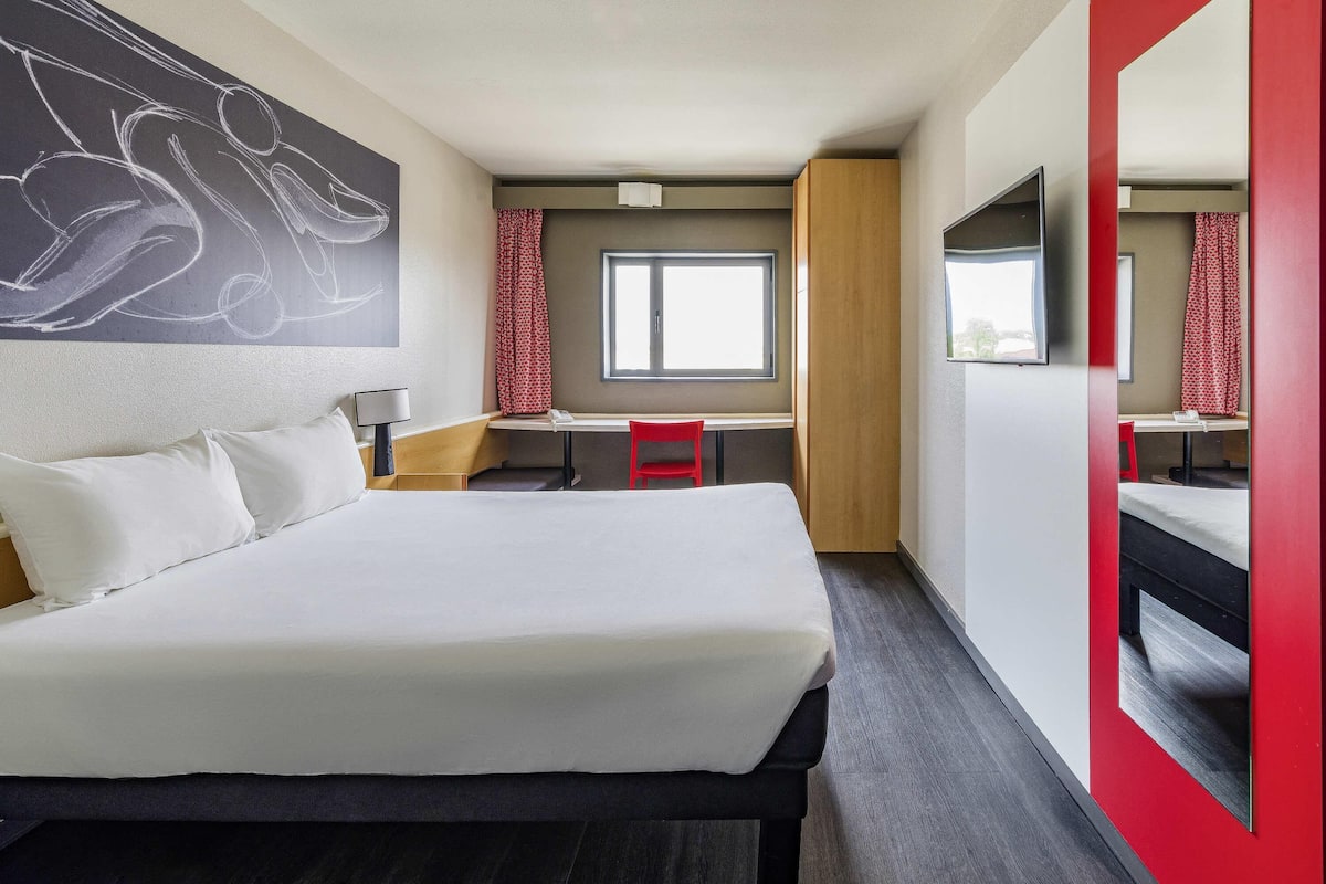 Une chambre d’hôtel avec un grand lit, un bureau, une chaise, une télévision et un mur d’accent de couleur rouge.