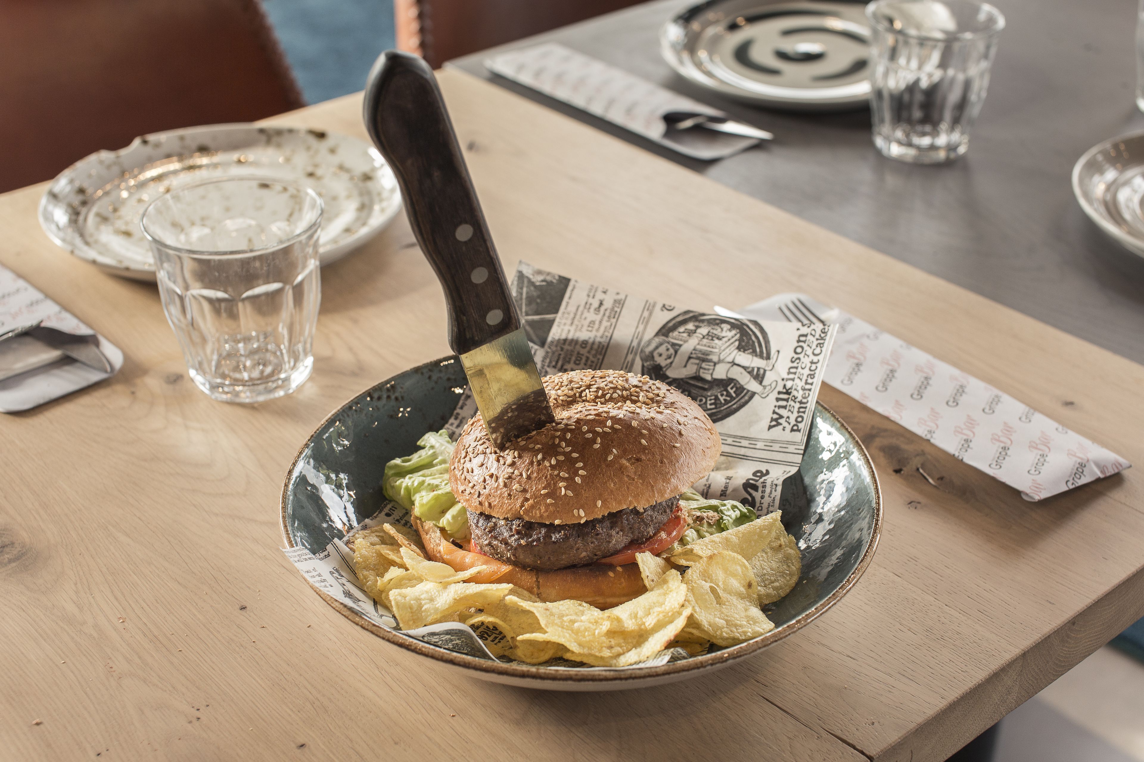 Hamburguesa amb un ganivet a sobre, damunt d'un plat de patates, sobre una taula de fusta.