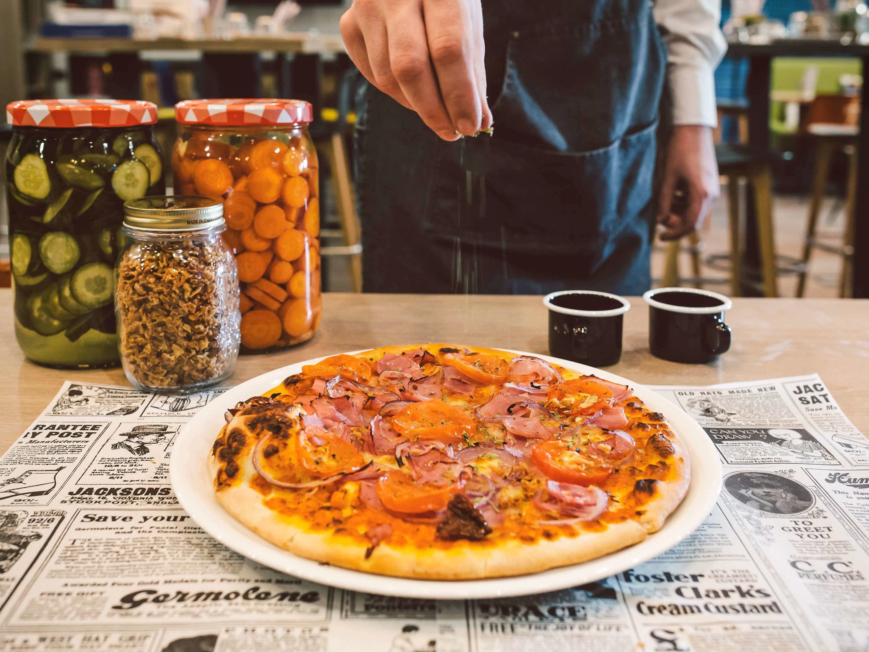Une pizza sur une assiette, garnie de divers ingrédients, un journal, et des bocaux de cornichons et de carottes.