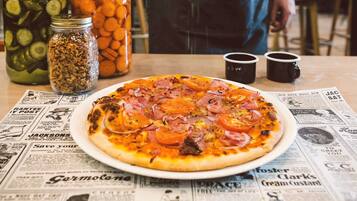 Une pizza sur une assiette, garnie de divers ingrédients, un journal, et des bocaux de cornichons et de carottes.