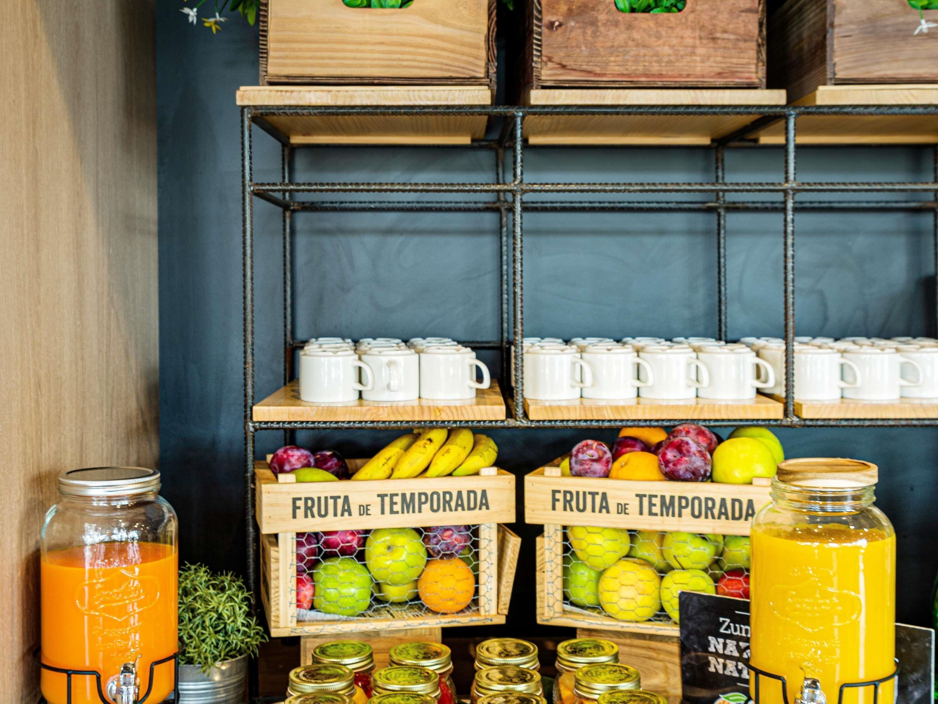 A kitchen area with wooden crates labeled 'FRUTA DE TEMPORADA' containing various fruits, a juice dispenser, and white mugs on shelves.