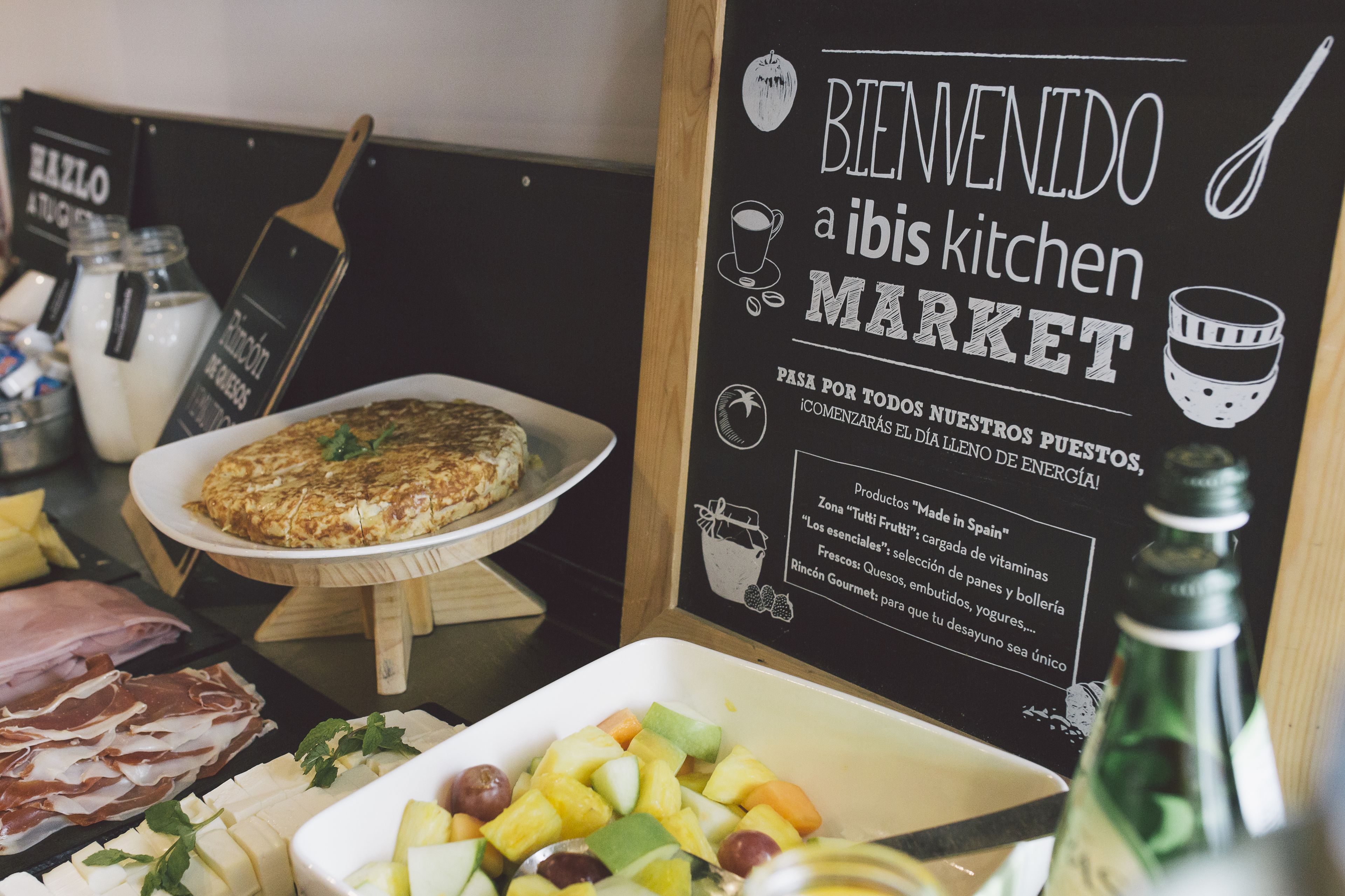 A breakfast buffet with a variety of foods including a quiche, fruit, and meats. A sign reads "Bienvenido a ibis kitchen Market".
