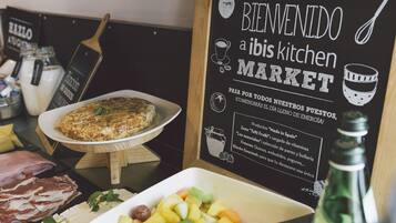 A breakfast buffet with a variety of foods including a quiche, fruit, and meats. A sign reads "Bienvenido a ibis kitchen Market".