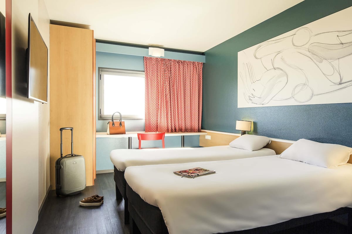 Une chambre d’hôtel avec un lit, un bureau, une chaise, une valise et une fenêtre avec des rideaux.