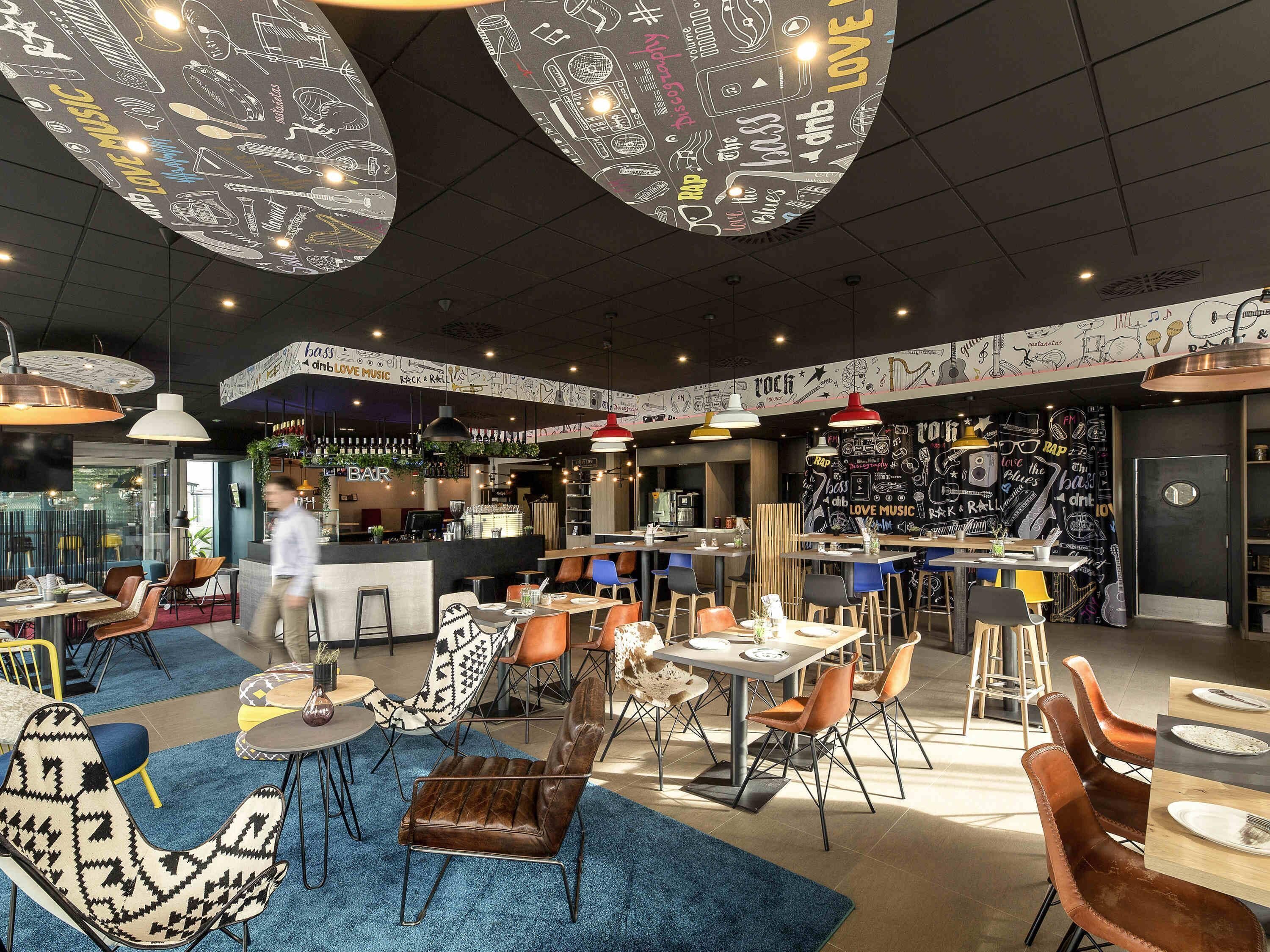 Een modern restaurant met een bar, verschillende zithoeken zoals tafels en stoelen, en een plafond met hangende lampen.