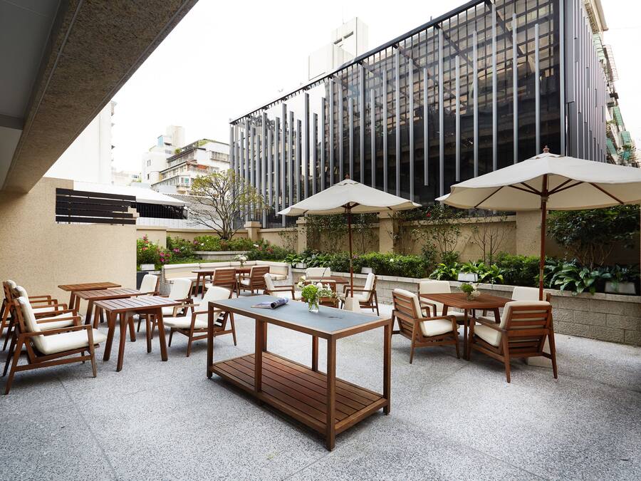 An outdoor patio area with wooden furniture and umbrellas, surrounded by greenery and a modern building in the background.