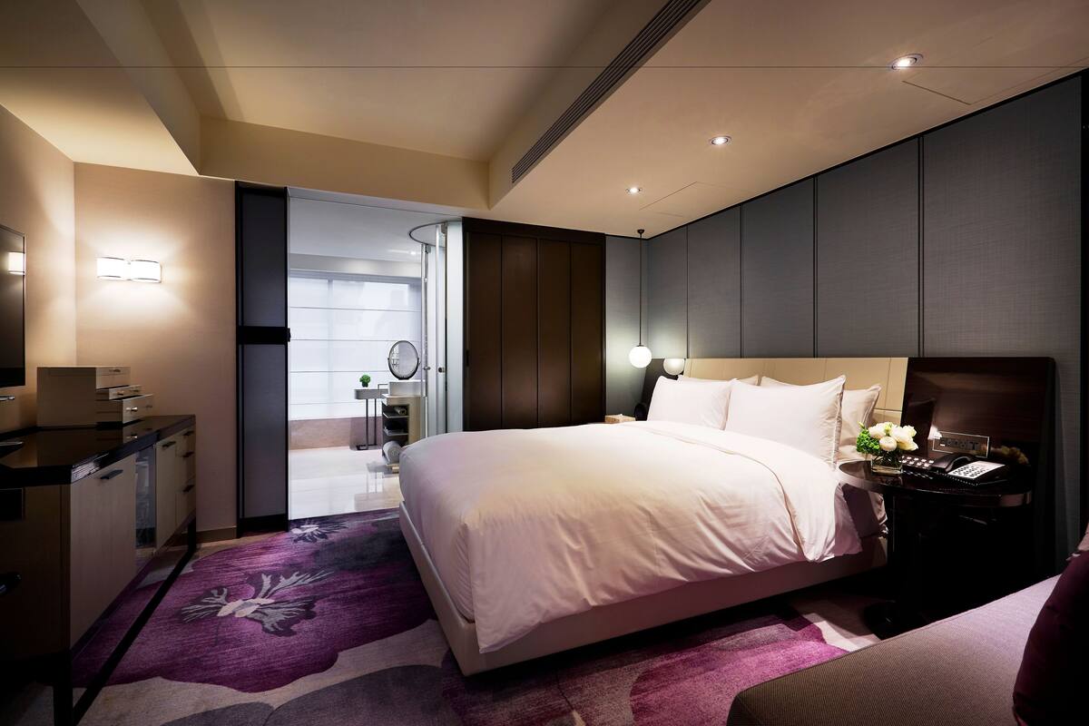 A modern hotel room with a large bed, bedside table with a lamp, and a desk with a chair.