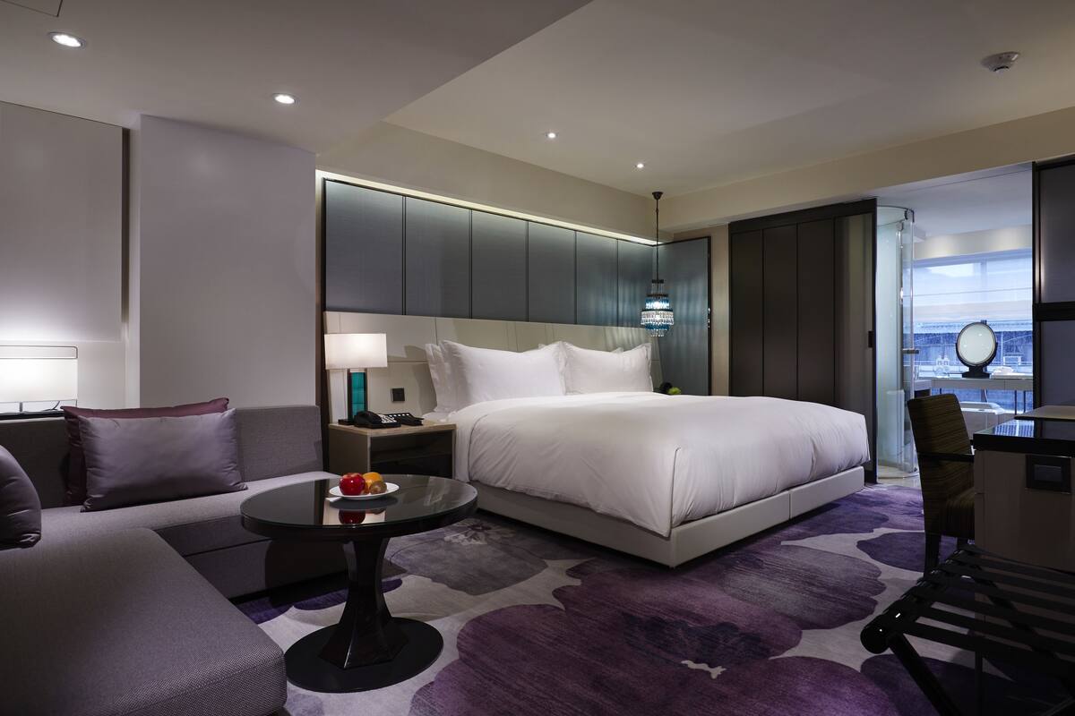 A modern hotel room with a large bed, a round table, a sofa, and a desk with a chair.