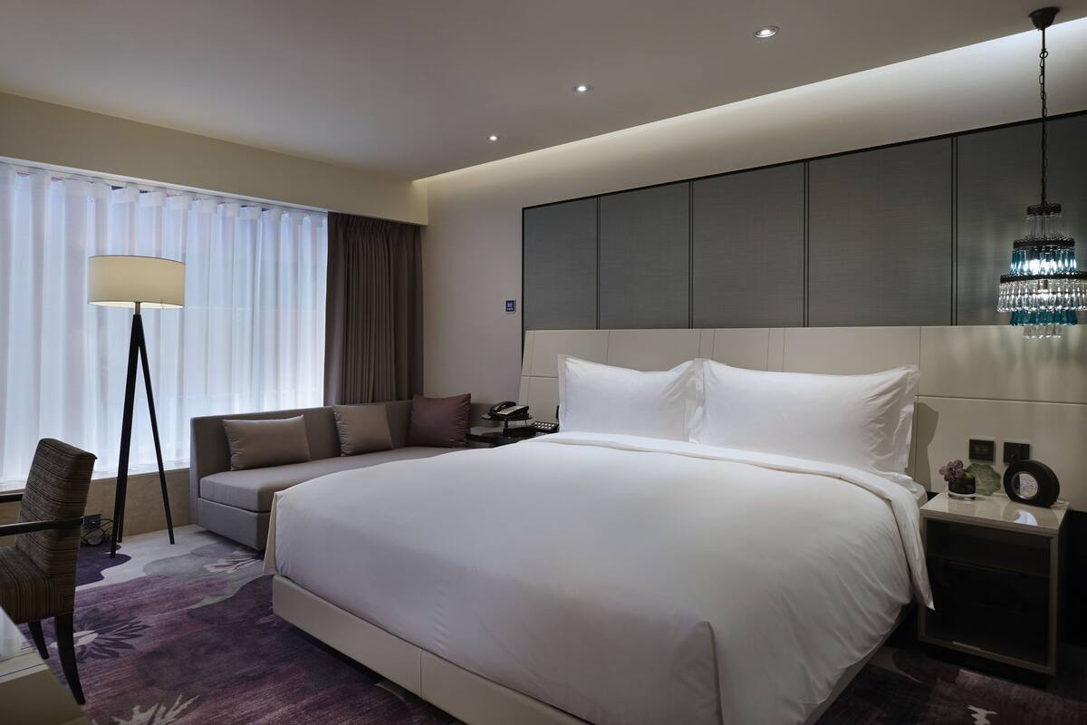 A modern hotel room with a large bed, a sofa, a desk with a lamp, and a chandelier.