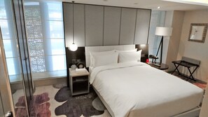 Une chambre d’hôtel moderne avec un grand lit, des tables de chevet, une petite table, une lampe et une vue à travers une fenêtre à motifs.