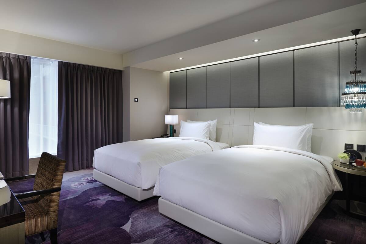 A hotel room with two beds, a desk, a chair, a lamp, and a chandelier.
