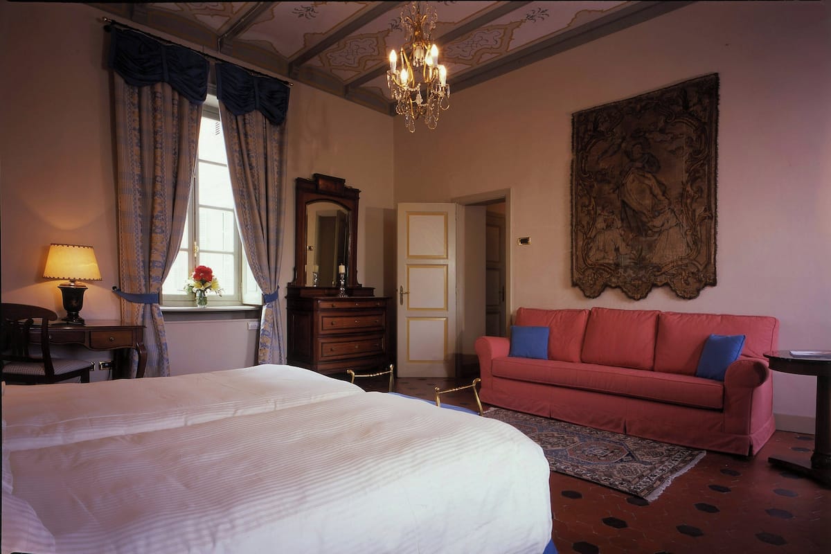 Ein Schlafzimmer mit einem großen Bett, einem roten Sofa, einer Kandelaberleuchte, einem großen Gemälde und einer Holzkommode.