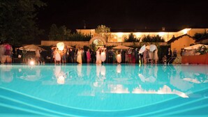 A nighttime poolside scene with a clear pool, a building with arches and a large wheel structure in the background, and people gathered around. - Sina Villa Matilde (Romano Canavese)