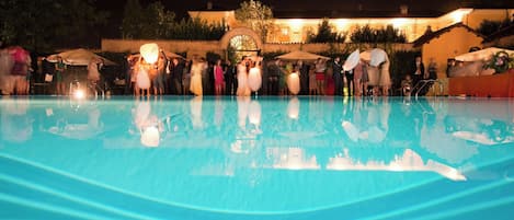 Escena nocturna junto a la piscina, con agua cristalina, un edificio con arcos y una gran estructura circular al fondo, y gente reunida.