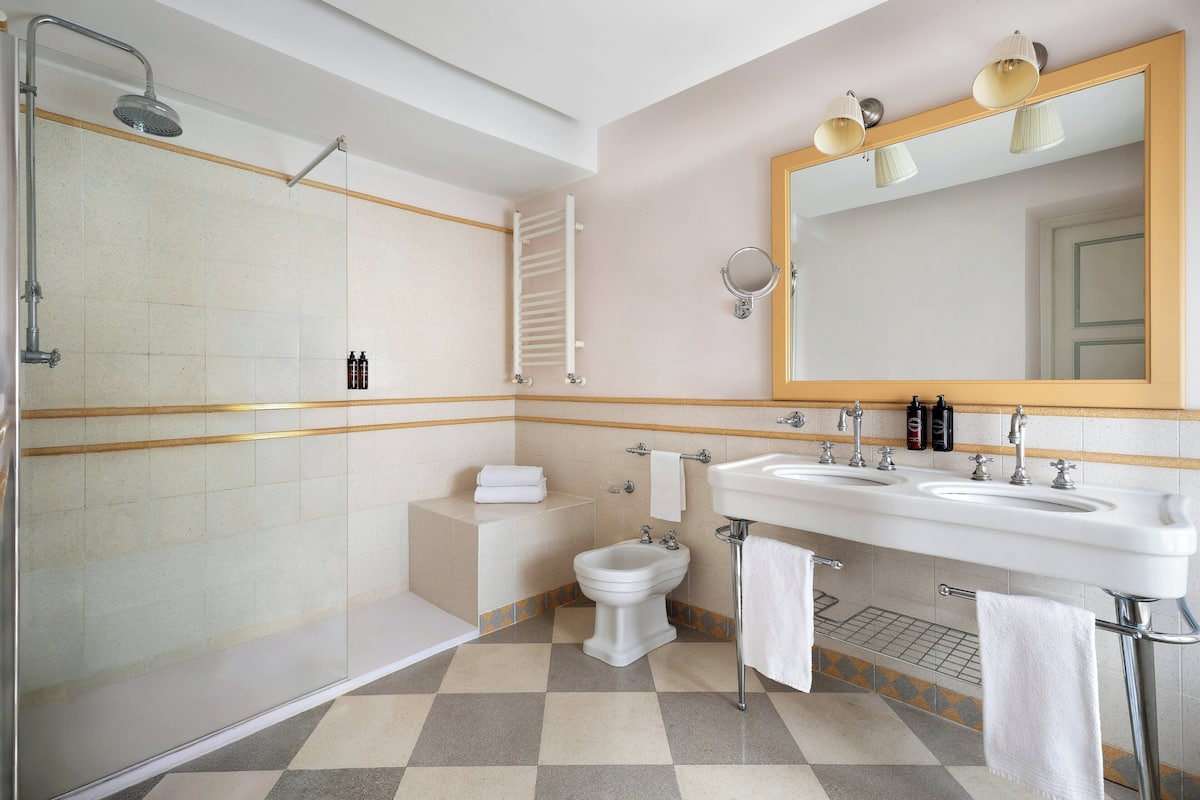 A bathroom with a shower, bidet, and two sinks with a mirror above.