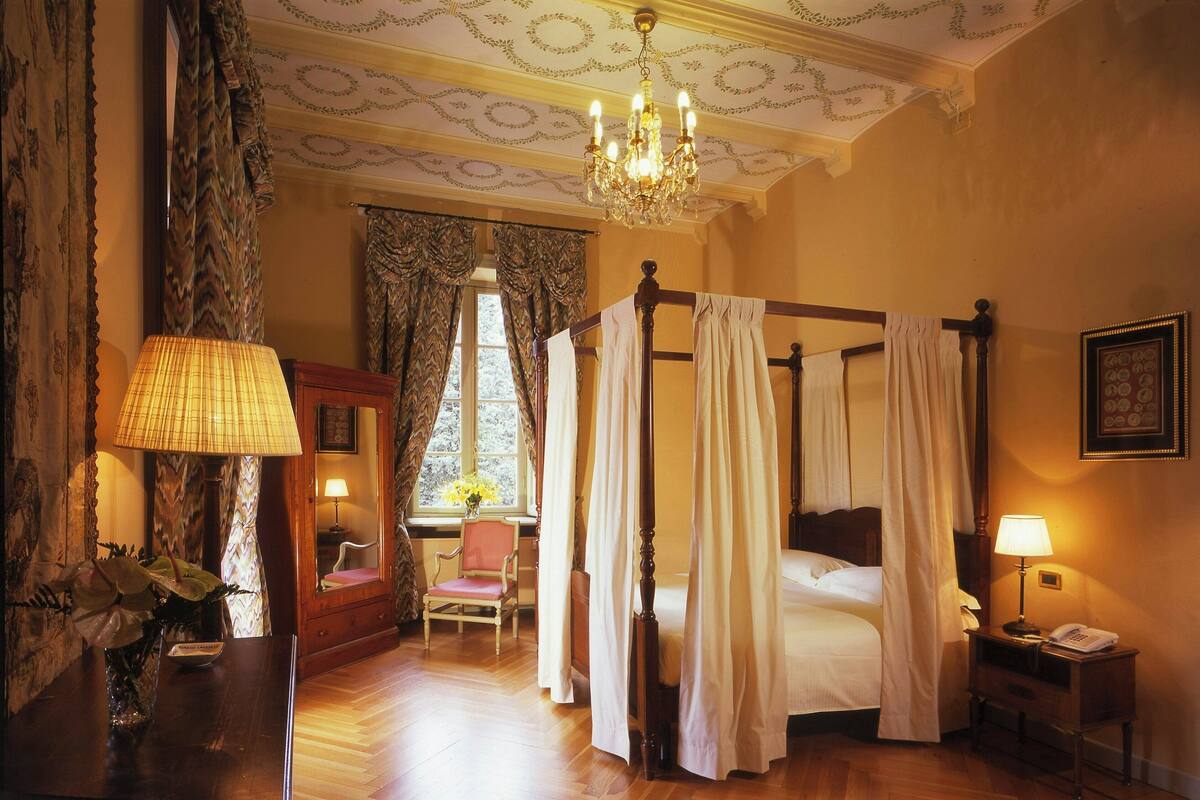 Ein Bett mit Baldachin, ein Sessel, ein Nachttisch mit Lampe und ein Fenster mit Vorhängen.